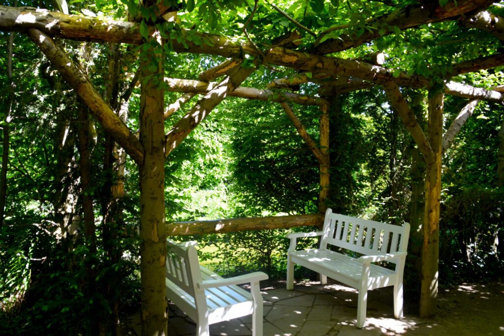 Weiße Parkbänke unter einem schattenspendenden Holzpavillon inmitten eines üppig bewachsenen Gartens, der zum Verweilen und Entspannen einlädt.