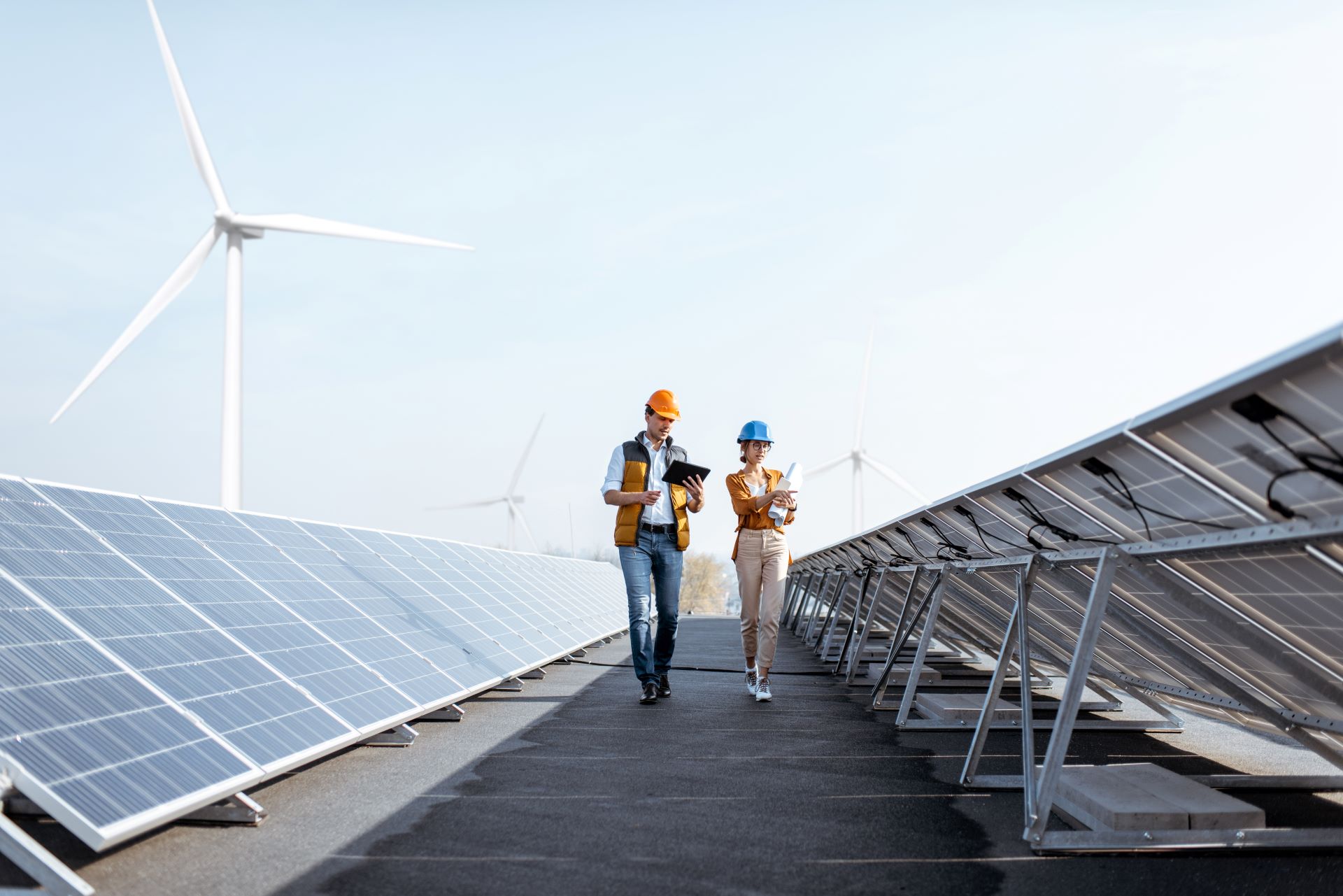 Zwei Ingenieure gehen entlang einer Solaranlage, im Hintergrund sind Windkraftanlagen zu sehen. Sie verwenden Tablets zur Überprüfung der Systeme