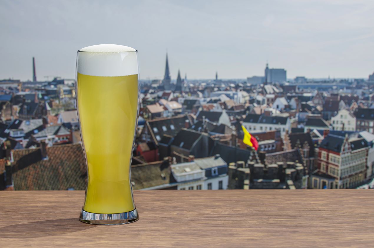 Ein Glas belgisches Bier, inspiriert von Tripel Karmeliet, steht auf einem Holztisch vor einer historischen Stadtansicht mit belgischer Flagge.